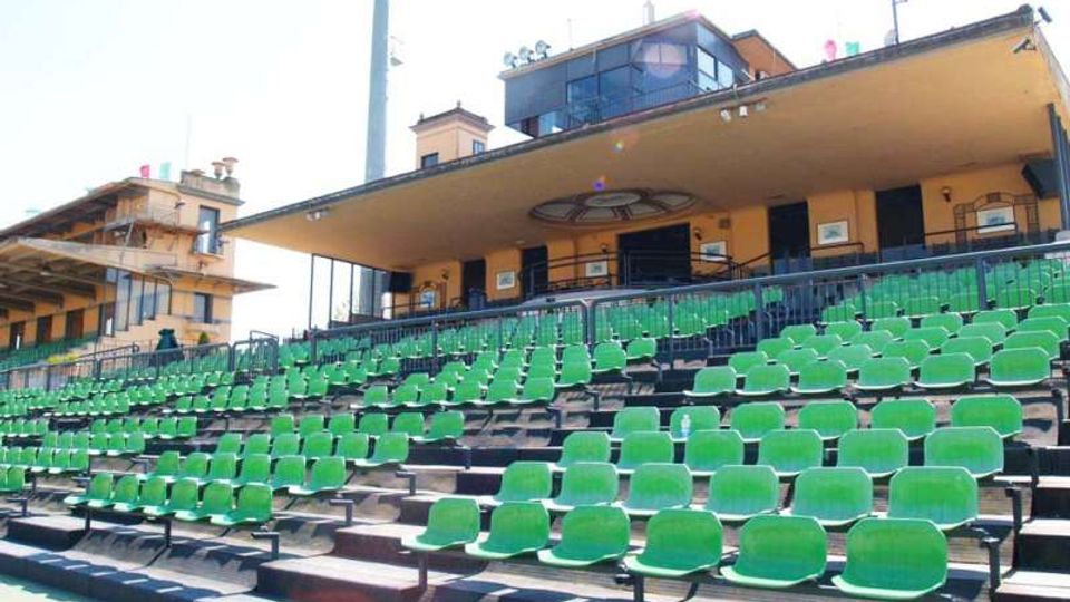 Gradinate di uno stadio con numerosi sedili verdi in file su livelli, delimitate da ringhiere metalliche. Sullo sfondo si vede la tribuna coperta con edificio in muratura gialla, finestre e struttura superiore vetrata.