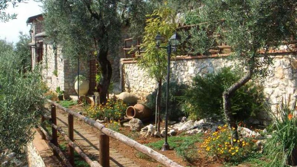 Vialetto in pietra con staccionata in legno che attraversa un giardino mediterraneo con ulivi, aiuole fiorite e grandi anfore in terracotta. Sullo sfondo si vede un muro in pietra con lampioni e un edificio rustico in pietra.
