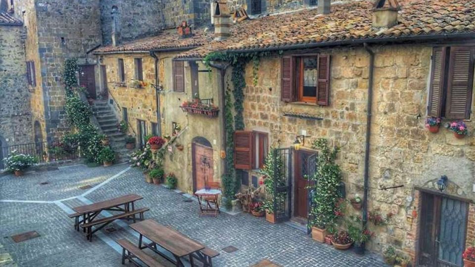 Piccola piazza lastricata in pietra con panche e tavoli in legno, circondata da case in muratura di pietra con tetti in coppi. Facciate con persiane in legno, portoni ad arco, scale esterne e numerosi vasi di fiori e piante rampicanti lungo i muri.