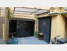 Cortile interno di un edificio con pavimento in cemento e pareti color ocra, coperto da una tettoia in metallo e pannelli traslucidi. Si vedono grandi portoni neri, tubazioni a vista, piante rampicanti e una piccola mensola in legno vicino all’ingresso.