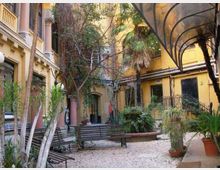 Cortile interno di un edificio con facciate gialle e balconi, pavimentato in ghiaia e arredato con panchine. Sono presenti molte piante e alberi in vaso e un’ampia tettoia trasparente ad arco lungo il lato destro.
