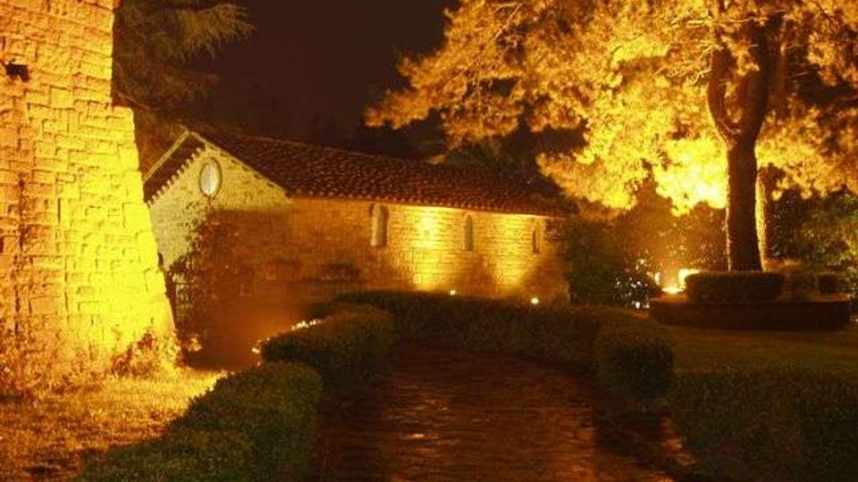 Percorso in pietra in un giardino con siepi basse, illuminato da luci notturne che evidenziano un edificio in pietra con tetto in tegole e una torre muraria a sinistra. Un grande albero sulla destra è illuminato dal basso, con sfondo di cielo scuro.