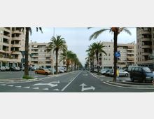 Viale urbano ampio con corsie e frecce direzionali sull’asfalto, fiancheggiato da palme e auto parcheggiate ai lati. Sullo sfondo si vedono palazzi residenziali moderni con balconi lungo entrambi i lati della strada.