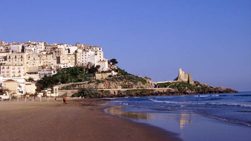 Veduta costiera con ampia spiaggia sabbiosa e mare calmo in primo piano, e un borgo di case bianche addossate a una collina rocciosa. Sulla punta del promontorio si distingue una torre di avvistamento in pietra affacciata sul mare.