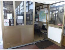 Ingresso di un piccolo ufficio o postazione di controllo con pareti vetrate e struttura metallica, porta aperta con cartello “SPINGERE”. All’interno si vedono sedie, una scrivania con attrezzature e una mappa appesa alla parete.