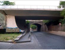 Sottopasso stradale urbano in cemento con carreggiata in sampietrini e marciapiede laterale, sotto un grande viadotto. Sulle pareti sono presenti graffiti e, in fondo, si vede un secondo arco in muratura con vegetazione ai lati.