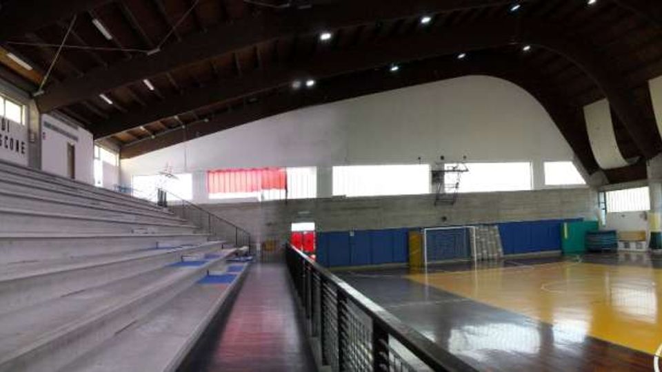 Interno di un palazzetto dello sport con parquet segnato per basket e pallamano, spalti in cemento su un lato e ringhiera lungo il corridoio. Tetto alto ad arco con travi a vista, grandi finestre e pareti con pannelli e striscioni.