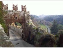 Porta fortificata in pietra con merli e grande arco che scavalca una strada stretta, affiancata da muri rocciosi. Sullo sfondo si vede una valle collinare con boschi e una strada che curva tra i pendii.