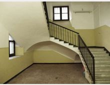 Interno di un vano scala in edificio, con pareti bicolore bianco e beige, pavimento in piastrelle e scalinata in pietra con ringhiera in ferro scuro. Una grande finestra quadrata e una piccola finestra laterale illuminano lo spazio, con un arco sotto la rampa principale.
