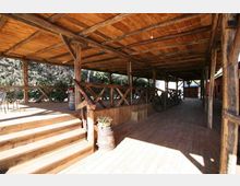 Portico rustico in legno con tetto a travi a vista e pavimento in assi, delimitato da una staccionata con montanti incrociati. A sinistra ci sono scalini e una pedana, con botti decorative e vista su alberi; il corridoio coperto si estende in profondità verso edifici sullo sfondo.