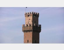 Torre in mattoni in stile medievale con sommità merlata e coronamento aggettante sostenuto da archetti, con piccole feritoie e finestre. La torre si staglia isolata contro un cielo azzurro.