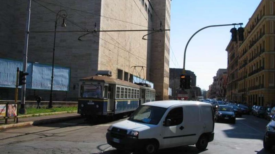 Strada urbana con binari del tram al centro, attraversata da una rete di cavi aerei e semafori. Lungo la via si vedono un grande edificio moderno in pietra e palazzi a più piani, con traffico di veicoli e un tram in corsa.