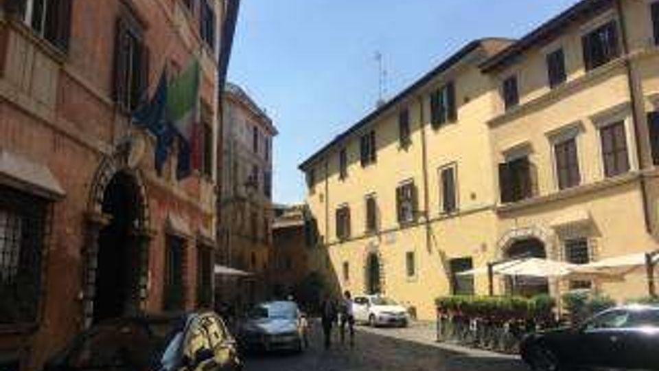 Strada urbana stretta con pavimentazione in ciottoli, fiancheggiata da edifici storici a più piani con facciate in intonaco e finestre con persiane. Auto parcheggiate lungo i lati e, sul lato destro, un’area con tavolini e ombrelloni davanti a un edificio.