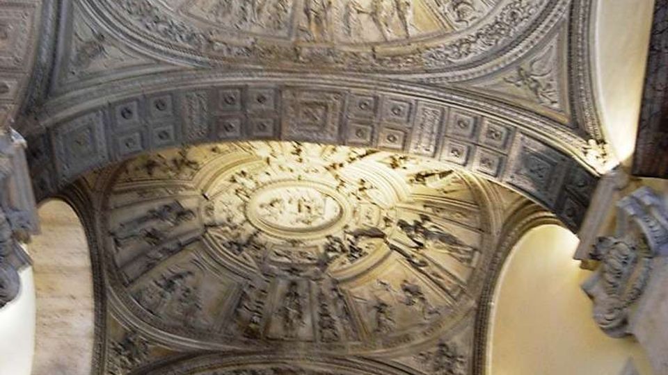 Interno di una chiesa o edificio storico con soffitto a volte decorate da stucchi in rilievo, medaglioni ovali e scene figurative. Archi e cornici ornate incorniciano la volta, con illuminazione calda che mette in risalto i dettagli.