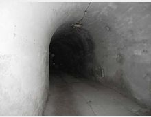 Lungo tunnel sotterraneo in cemento con soffitto a volta e pareti grezze, illuminato debolmente da una lampada a soffitto. Il pavimento è in lastre e il corridoio si restringe verso un’area più buia in fondo.