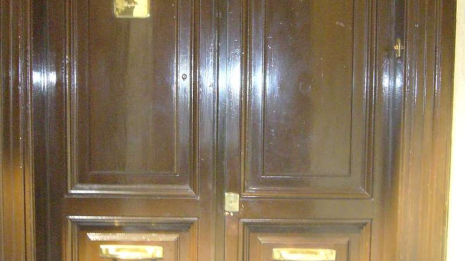 Portone d’ingresso a due ante in legno scuro lucido, con riquadri in rilievo e ferramenta in ottone. Sono visibili due fessure per la posta e una piccola targhetta sul battente sinistro.
