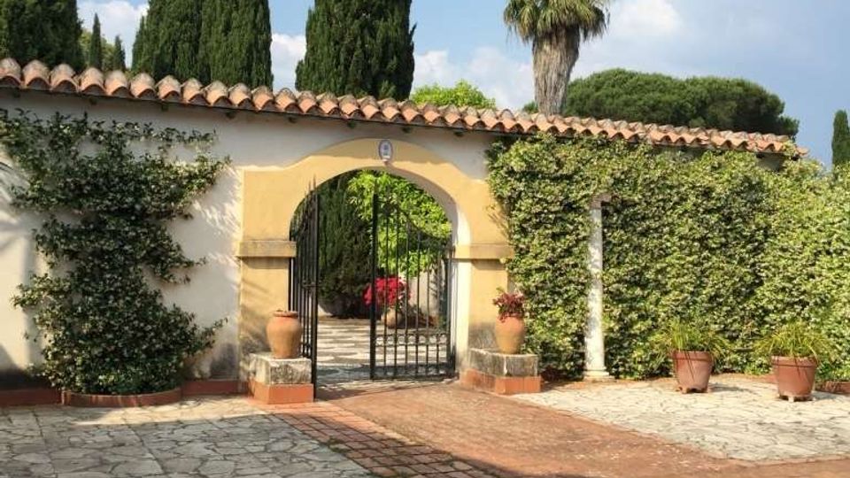 Cortile pavimentato in pietra con una parete intonacata e tetto in coppi, parzialmente coperta da edera, con un arco centrale che incornicia un cancello in ferro battuto. Sullo sfondo si vedono alti cipressi e una palma sotto un cielo azzurro con nuvole.