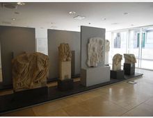 Interno di una sala museale moderna con pavimento in pietra chiara, pareti espositive grigie e illuminazione a soffitto, con rilievi e frammenti scultorei in pietra su piedistalli. Grandi finestre sul lato destro lasciano entrare luce naturale e mostrano edifici all’esterno.