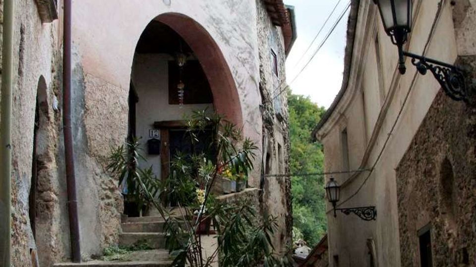 Vicolo stretto in un borgo con muri in pietra e intonaco, una scalinata che sale verso un grande arco d’ingresso e vasi di piante lungo i gradini. Sulla facciata opposta si vedono lampioni in ferro battuto e cavi sospesi, con vegetazione sullo sfondo tra gli edifici.