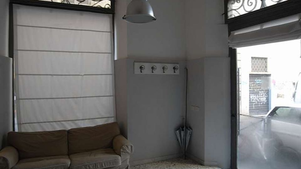 Interno di un piccolo locale con pavimento in graniglia, pareti bicolore e lampada a sospensione. A sinistra c’è un divano sotto una finestra con tendina e grata decorativa, mentre a destra una grande vetrata/porta su strada con tenda e ombrello nell’angolo.