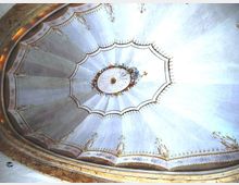 Vista verso l’alto di una cupola interna decorata, con pannelli radiali azzurro chiaro e motivi ornamentali dorati lungo il bordo. Al centro si vede un medaglione ovale con ghirlanda floreale e rosetta, circondato da cornici modanate.