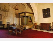 Interno di una stanza storica con soffitto a volte e pareti in pietra intonacata, arredata con un grande letto a baldacchino, tavolo e sedie in legno. Nella sala si vedono anche un cassettone, un ritratto appeso alla parete e una lanterna sospesa al soffitto su pavimento rivestito da tappeto rosso.