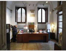 Interno di una biglietteria o reception in un edificio storico, con grande banco in legno al centro e materiali informativi esposti. Sullo sfondo ci sono due alte finestre ad arco con inferriate, pareti bianche con illuminazione a binario e una statua in una nicchia accanto a una porta vetrata.