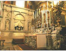 Interno di una chiesa con altare barocco riccamente decorato, candelabri alti e pannelli dorati, affiancato da una parete in pietra con nicchie e statue. Il soffitto e le pareti superiori sono ornati da dipinti e cornici decorative.