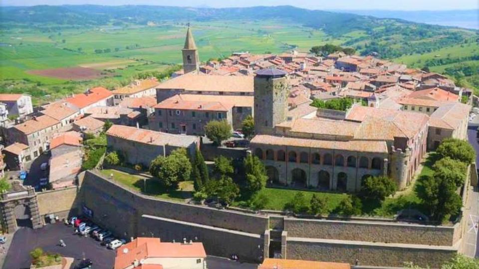 Veduta aerea di un borgo storico con edifici in pietra e tetti in coppi, dominato da un complesso fortificato con mura perimetrali, torre quadrata e chiesa con campanile. Sullo sfondo si estendono colline e campi coltivati.