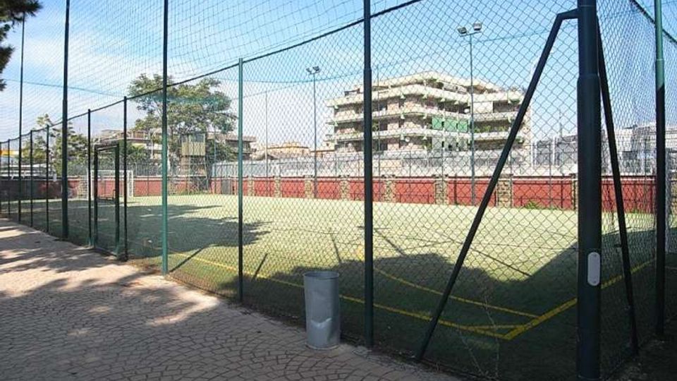 Campo sportivo all’aperto con erba sintetica, recinzione alta in rete metallica e fari per l’illuminazione. Accanto c’è un vialetto pavimentato e sullo sfondo si vedono palazzi residenziali e alcuni alberi.