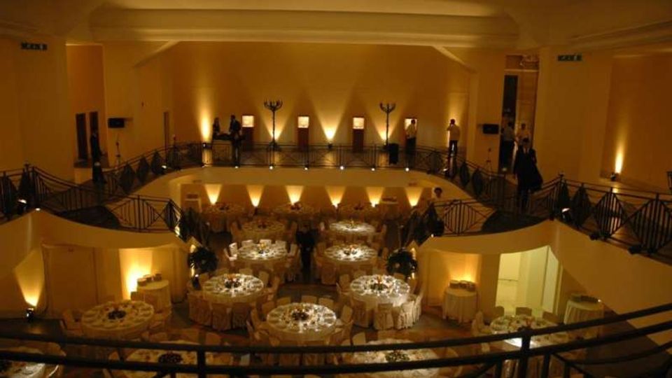 Ampia sala per ricevimenti su due livelli con soppalco e ringhiere in ferro, illuminata da luci calde a parete. Al piano inferiore sono disposti numerosi tavoli rotondi apparecchiati con tovaglie chiare e sedie, con scala laterale e passaggi lungo il perimetro.