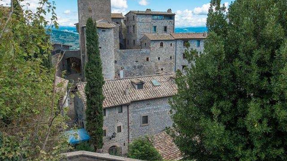 Scalinata in pietra con muretto curvo che scende verso un borgo medievale in muratura, con edifici dai tetti in coppi e una torre rettangolare alta. Vegetazione ai lati della scala e panorama di colline sullo sfondo sotto un cielo con nuvole.