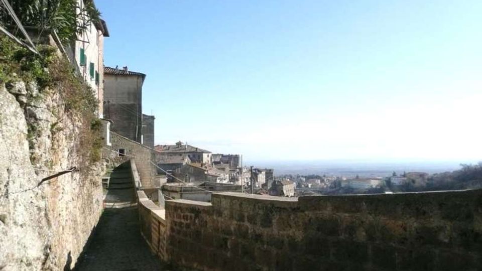 Stradina in salita lastricata lungo una parete rocciosa e case in pietra, affiancata da un muretto di contenimento. Sullo sfondo si vede un borgo collinare e un ampio panorama di campagna fino all’orizzonte sotto un cielo limpido.
