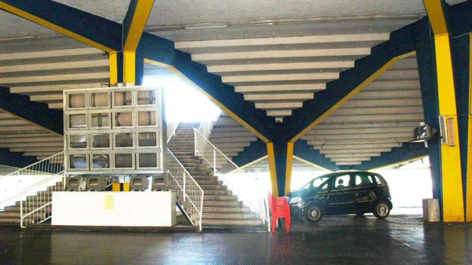 Interno di un parcheggio multipiano con ampie rampe e scale in cemento, travi strutturali verniciate in blu e giallo e pavimentazione scura. Al centro si vede una piccola area con gradinate e una struttura a finestrelle, con un’auto parcheggiata sul lato destro.