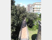 Passerella pedonale stretta in mattoni a spina di pesce con parapetti in muratura, immersa tra alberi e vegetazione e sospesa sopra un’area verde. Sullo sfondo si vedono palazzi residenziali moderni e una strada urbana.