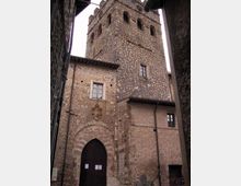 Torre medievale in pietra con finestre ad arco nella parte alta, vista dal basso in un vicolo stretto tra edifici in muratura. Alla base si vede un grande portale ad arco con stemma sopra e piccole finestre incorniciate.