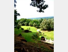 Vista dall’alto di un ampio parco collinare con prato verde, alberi folti e un sentiero chiaro che attraversa l’area. In primo piano si vede una piccola terrazza pavimentata con ombrelloni e pergola, mentre sullo sfondo si apre una pianura con campi e boschi all’orizzonte.