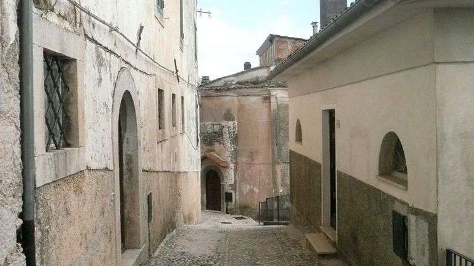 Vicolo stretto in pietra con pavimentazione a ciottoli, fiancheggiato da vecchi edifici in intonaco con porte ad arco e finestre con persiane. La strada in lieve discesa conduce verso altre case addossate, sotto un cielo parzialmente nuvoloso.