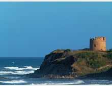 Torre costiera in pietra su un promontorio erboso affacciato sul mare, con scogliera e onde che si infrangono sulla riva sottostante. Il cielo sereno e l’orizzonte marino incorniciano la struttura isolata.
