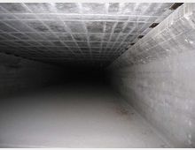 Interno di un lungo tunnel sotterraneo in cemento, con soffitto a griglia e pareti lisce che convergono verso un punto oscuro in lontananza. Il pavimento è spoglio e leggermente polveroso, illuminato debolmente.