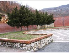Cortile pavimentato con aiuole rialzate delimitate da muretti in pietra e bordo in mattoni, con una fila di alberi sempreverdi davanti a un alto muro in mattoni. Sullo sfondo si vedono colline boscose.
