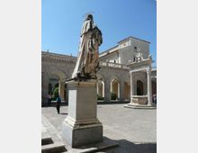 Cortile in pietra di un complesso religioso con portici ad archi e balaustre, con una grande statua su piedistallo in primo piano. Sullo sfondo si vede l’edificio in muratura con tetto a falde e una fontana in pietra.