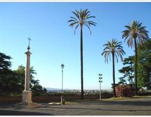 Belvedere con piazzale lastricato e vista panoramica sulla città in lontananza, delimitato da un muretto basso. In primo piano ci sono alte palme, un lampione e una colonna in pietra sormontata da una croce.