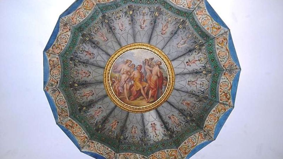 Affresco su soffitto circolare decorato con un motivo a raggiera e un medaglione centrale con figure classiche, incorniciato da bordure ornate in verde, oro e azzurro. Sfondo chiaro che evidenzia la struttura ottagonale della cornice esterna.
