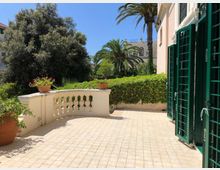Terrazza pavimentata con ringhiera in muratura e vasi di fiori, affacciata su un giardino con siepi e palme sotto cielo sereno. A destra si vede la facciata dell’edificio con grandi porte-finestre dotate di inferriate verdi.