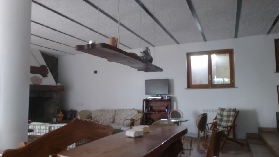 Soggiorno rustico con grande tavolo in legno e panche, soffitto alto con travi scure a vista e mensola sospesa. Sullo sfondo si vedono un camino ad angolo, una zona divani e sedie, una TV su mobile e una finestra rettangolare con cornice in legno.