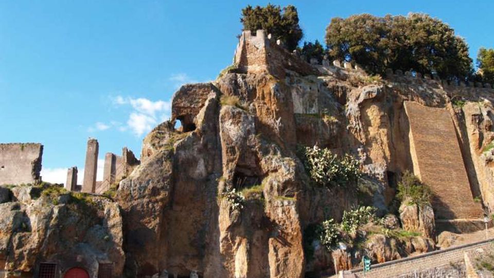 Rupe rocciosa con pareti di tufo erose e resti di mura e strutture in pietra sulla sommità, con alberi e vegetazione che crescono tra le fessure. In basso si vede una strada ai piedi della roccia e un cielo sereno sullo sfondo.