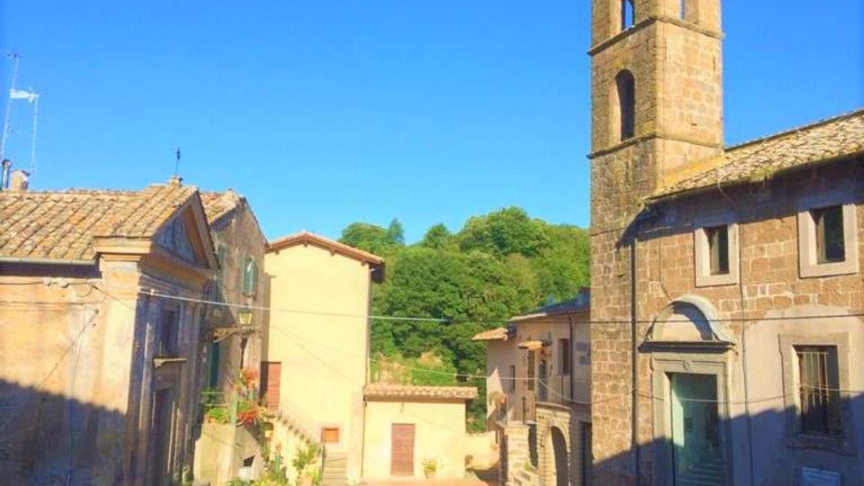 Piazzetta di un borgo con case in pietra e intonaco e una chiesa in muratura con campanile alto e aperture ad arco. Sullo sfondo si vedono colline alberate e cielo sereno, con scalinate e stradine lastricate che convergono nella piazza.