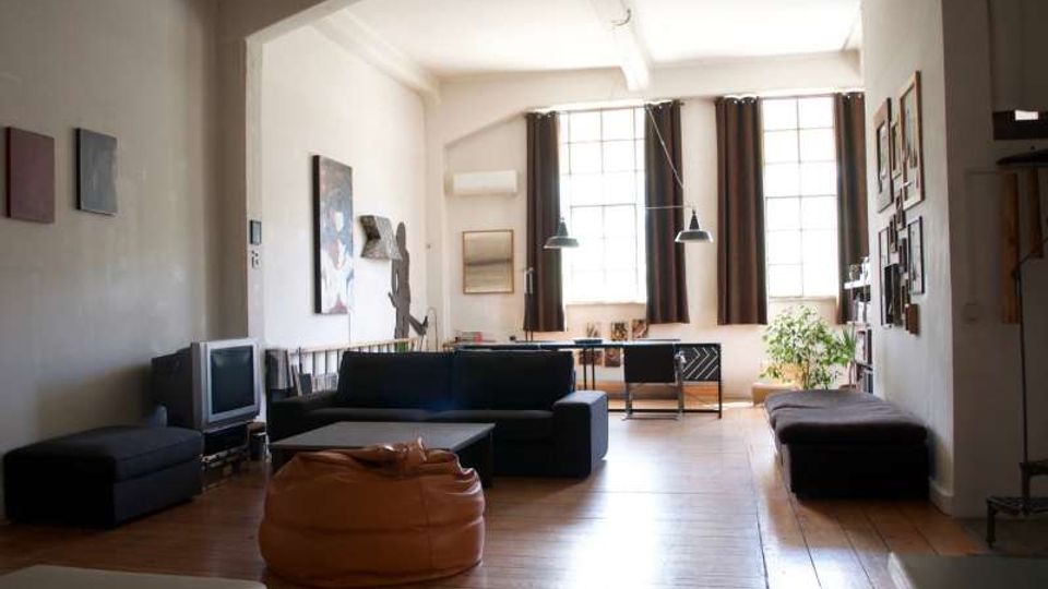 Ampio soggiorno in stile loft con pavimento in legno, divano scuro e pouf, pareti con quadri e lampade a sospensione. Sul fondo grandi finestre a riquadri con tende scure illuminano la stanza, con una piccola area studio e una pianta vicino alla finestra.