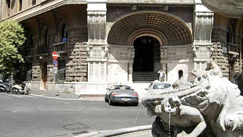 Ingresso di un edificio storico in pietra con grande arco decorato e scalinate, affacciato su una strada urbana. In primo piano si vede una fontana monumentale in pietra con sculture e vasca circolare.